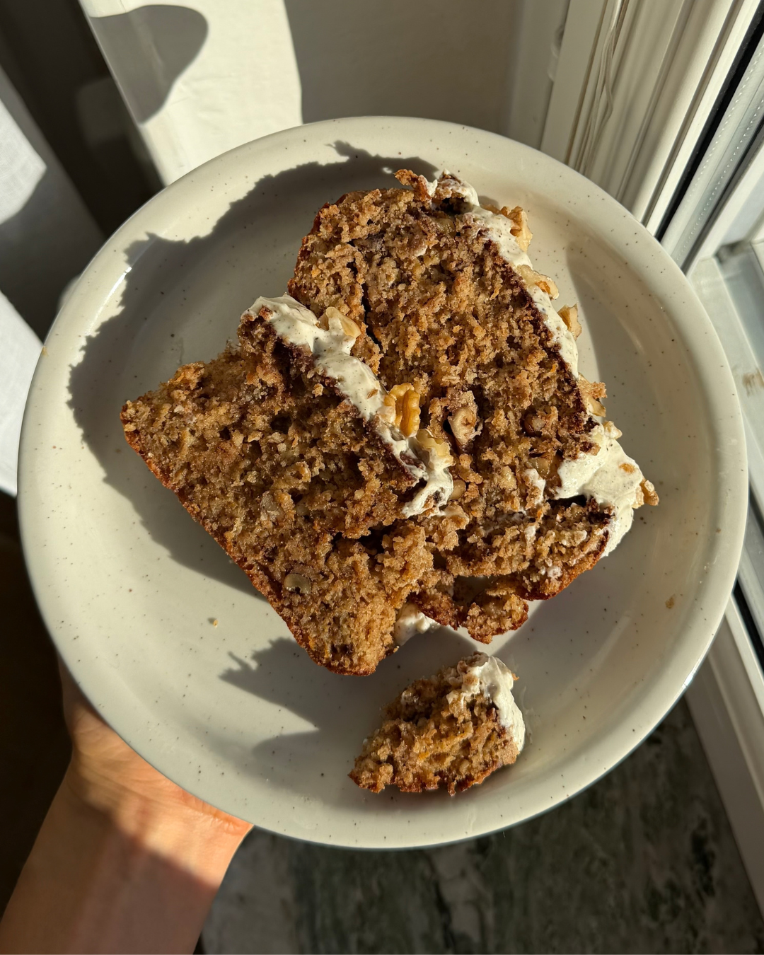 Vitt fat med två skivor av en saftig glutenfri morotskaka utan vitt socker med frosting och valnötter på toppen