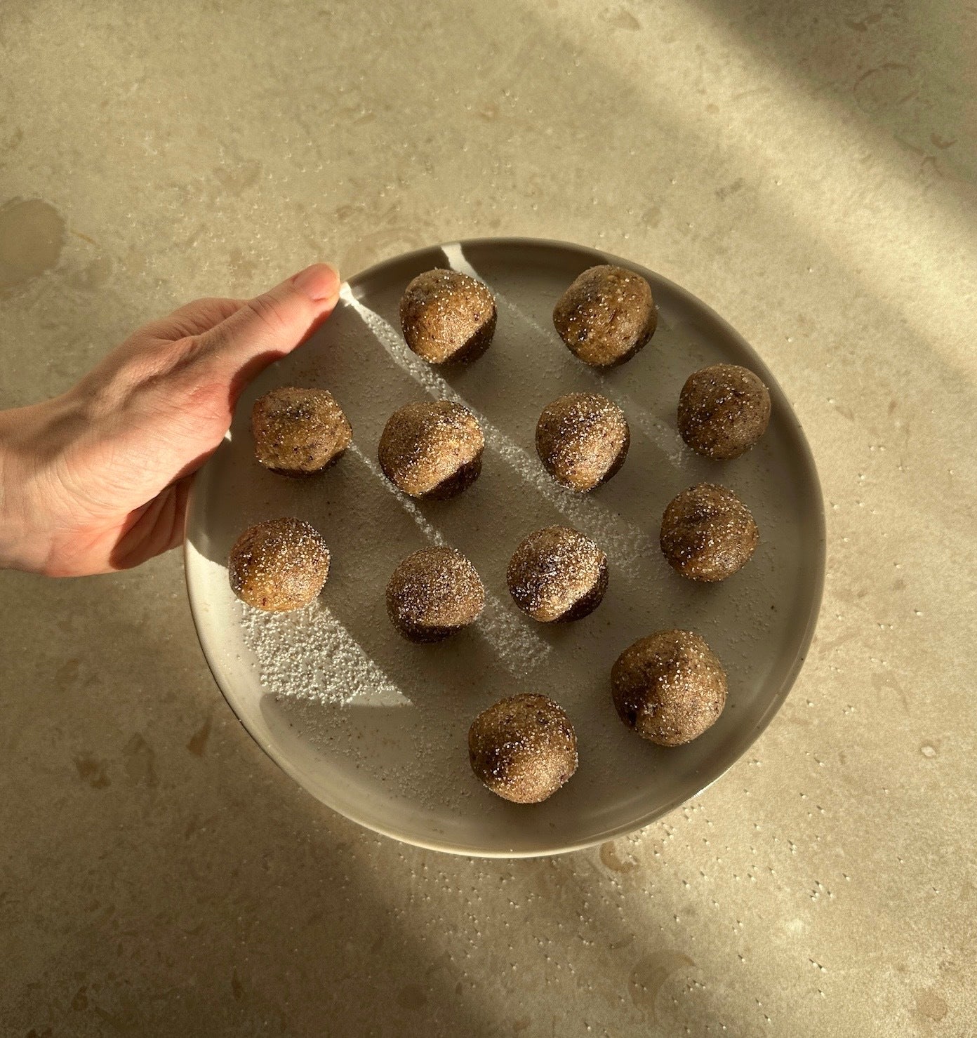 Hand som håller vitt fat i mjukt naturligt ljus med sockerfria rawfoodbollar med smak av kanelbulle på